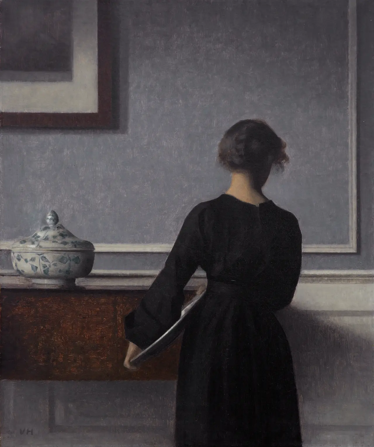 A woman in a black dress stands with her back turned, holding a large reflective plate beside dark furniture and a blue-and-white covered vessel.